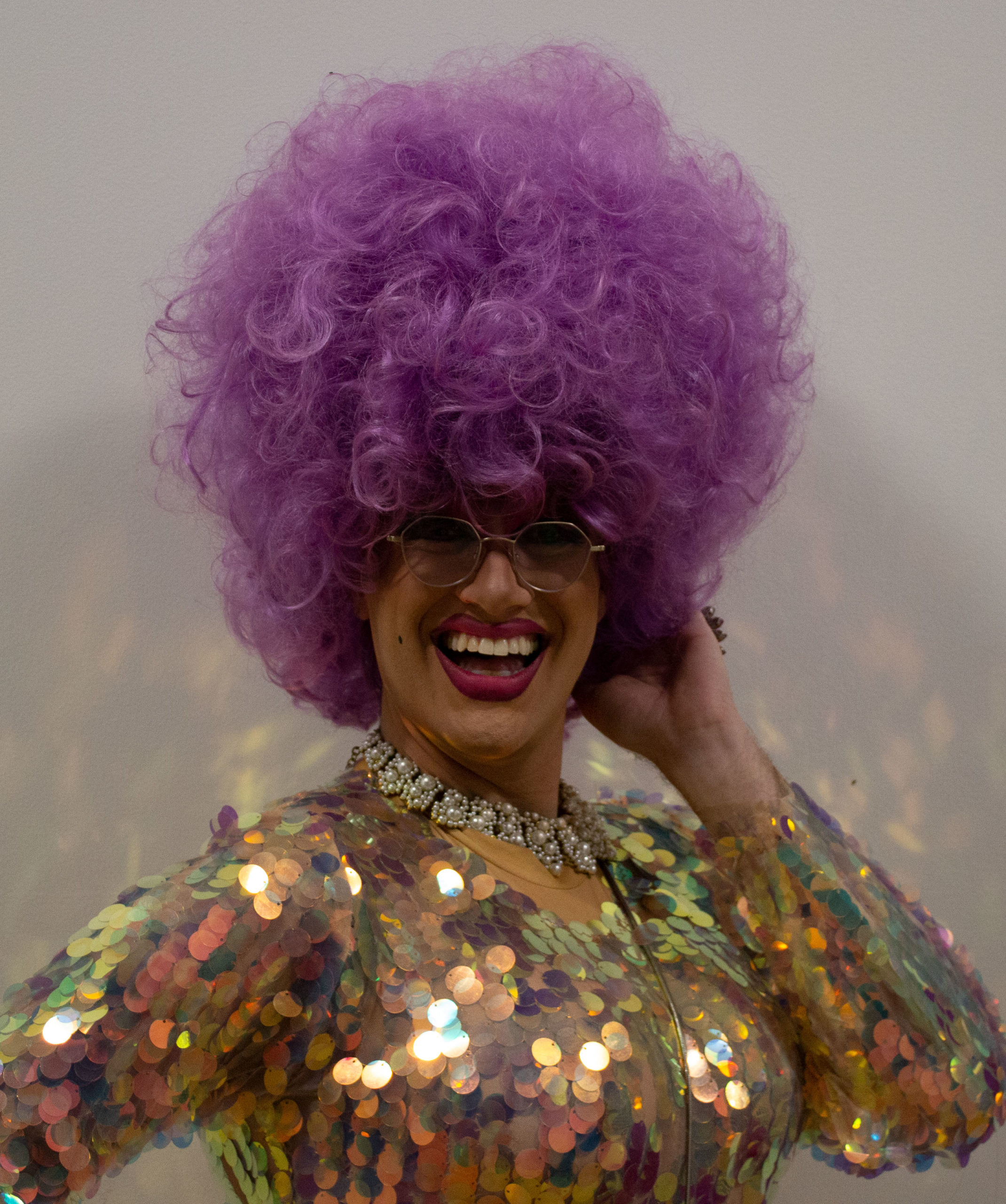 Pictured is Isaac Simmons in drag as Ms. Penny Cost. They are wearing a shiny dress with scales and a pink/purple wig.