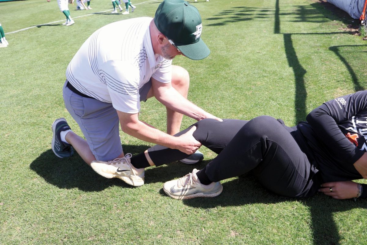 Head Athletic Trainer Dreher assists a student-athlete with a lower-leg stretch during practice, focusing on injury prevention and recovery
